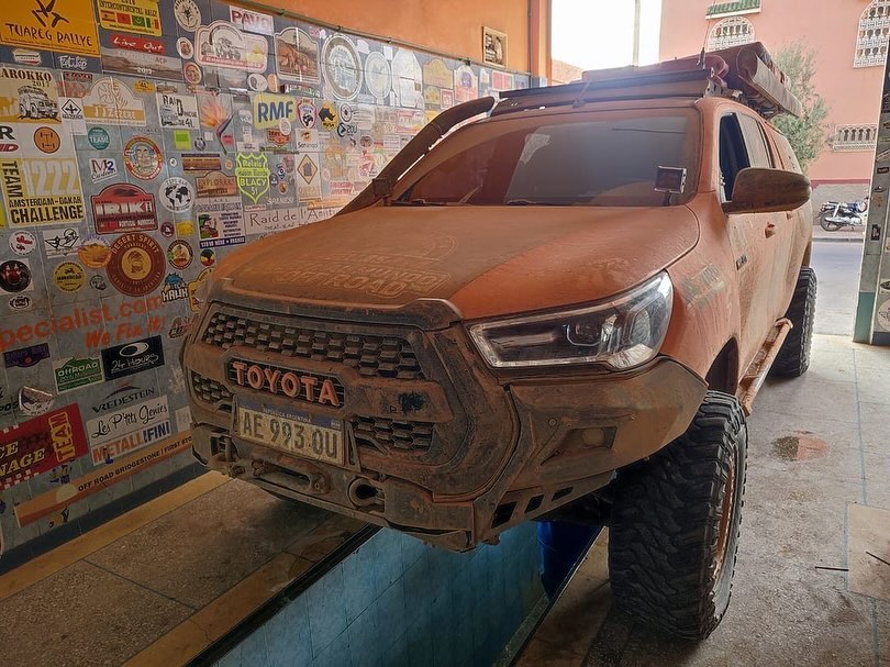 Dust-covered orange Toyota Hilux with Argentinian plates on the workshop ramp at Garage Iriki after crossing the desert