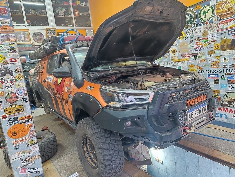Orange Toyota Hilux of the Argentinian SinHuellas 4x4 team with the hood open at Garage Iriki Zagora, sticker wall in the background