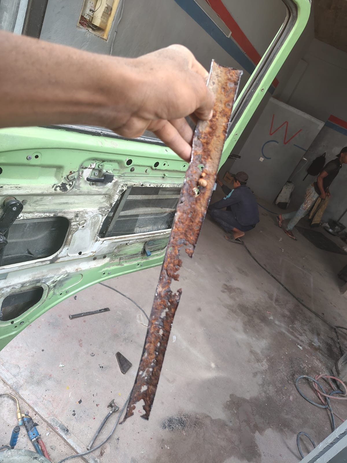 Mechanic holding up a heavily rusted metal strip cut out of the VW camper door frame at Garage Iriki