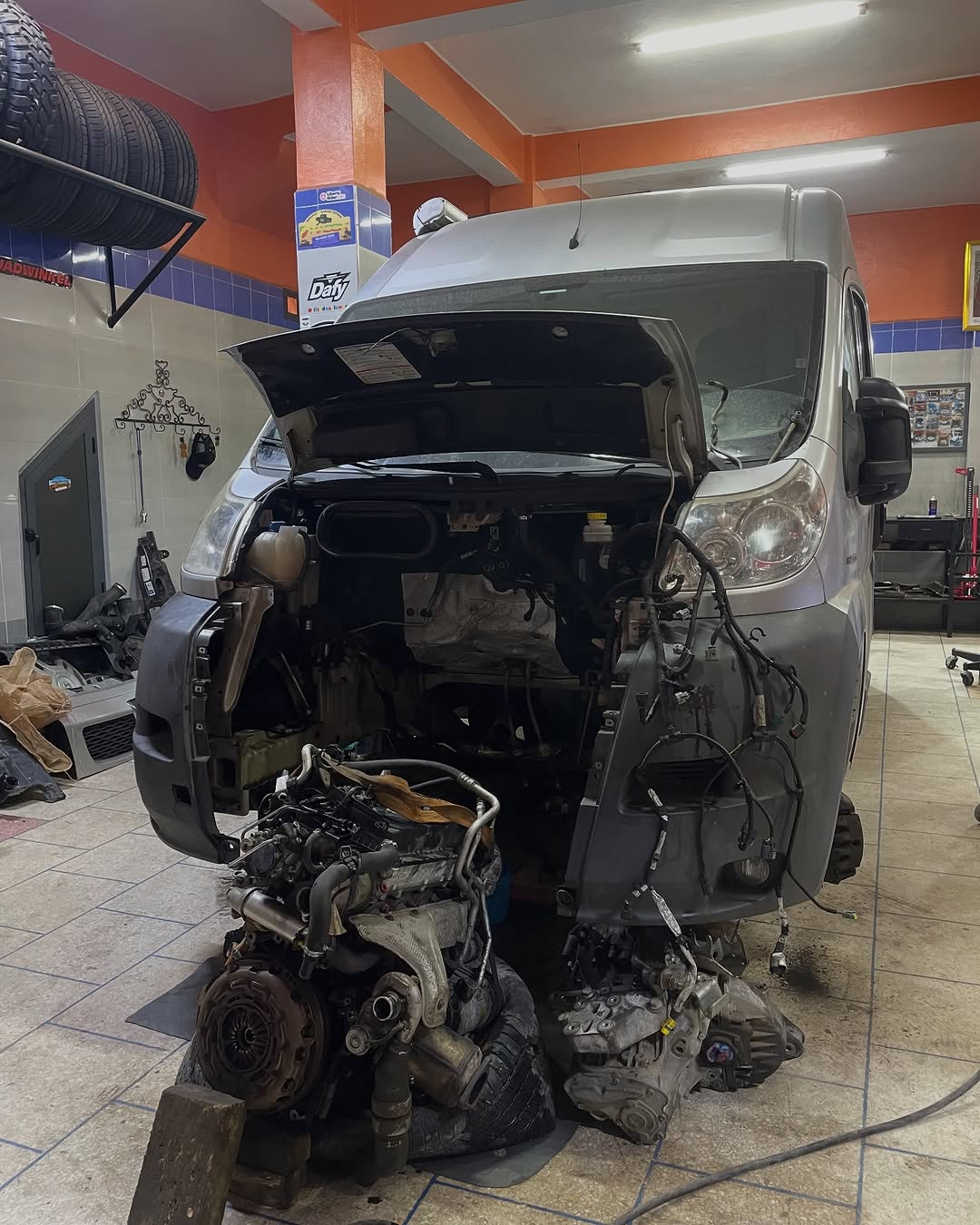 Front end of the silver Peugeot Boxer camper stripped down with the removed engine and transmission on the workshop floor