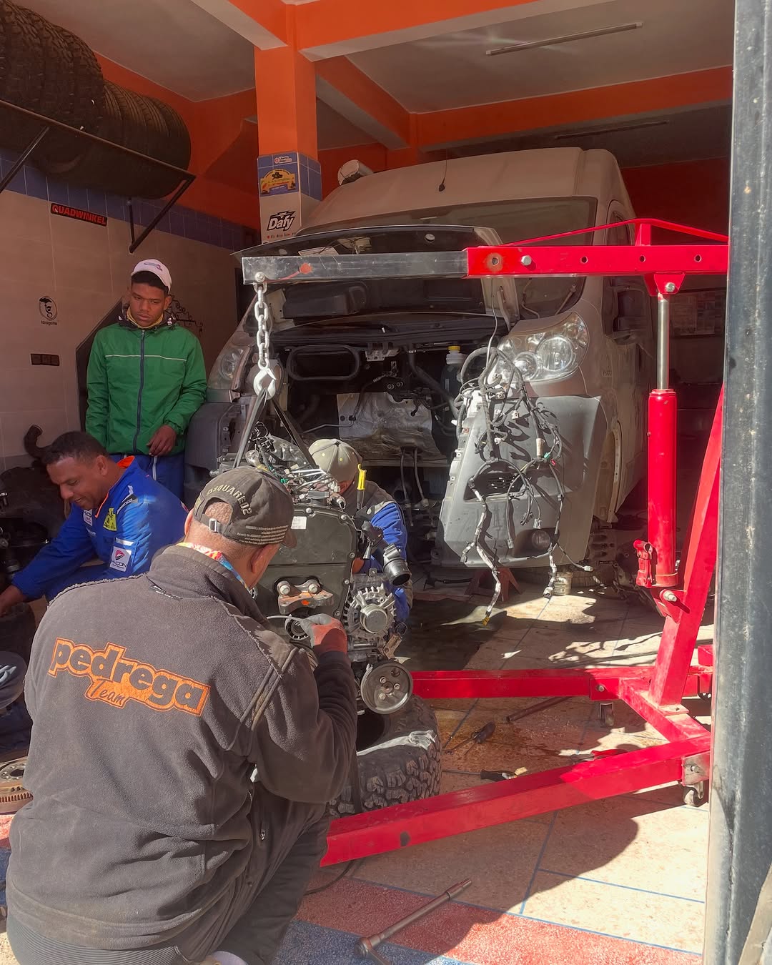 Mechanics using a red engine crane to lift a rebuilt engine into the silver Peugeot Boxer at Garage Iriki, Zagora