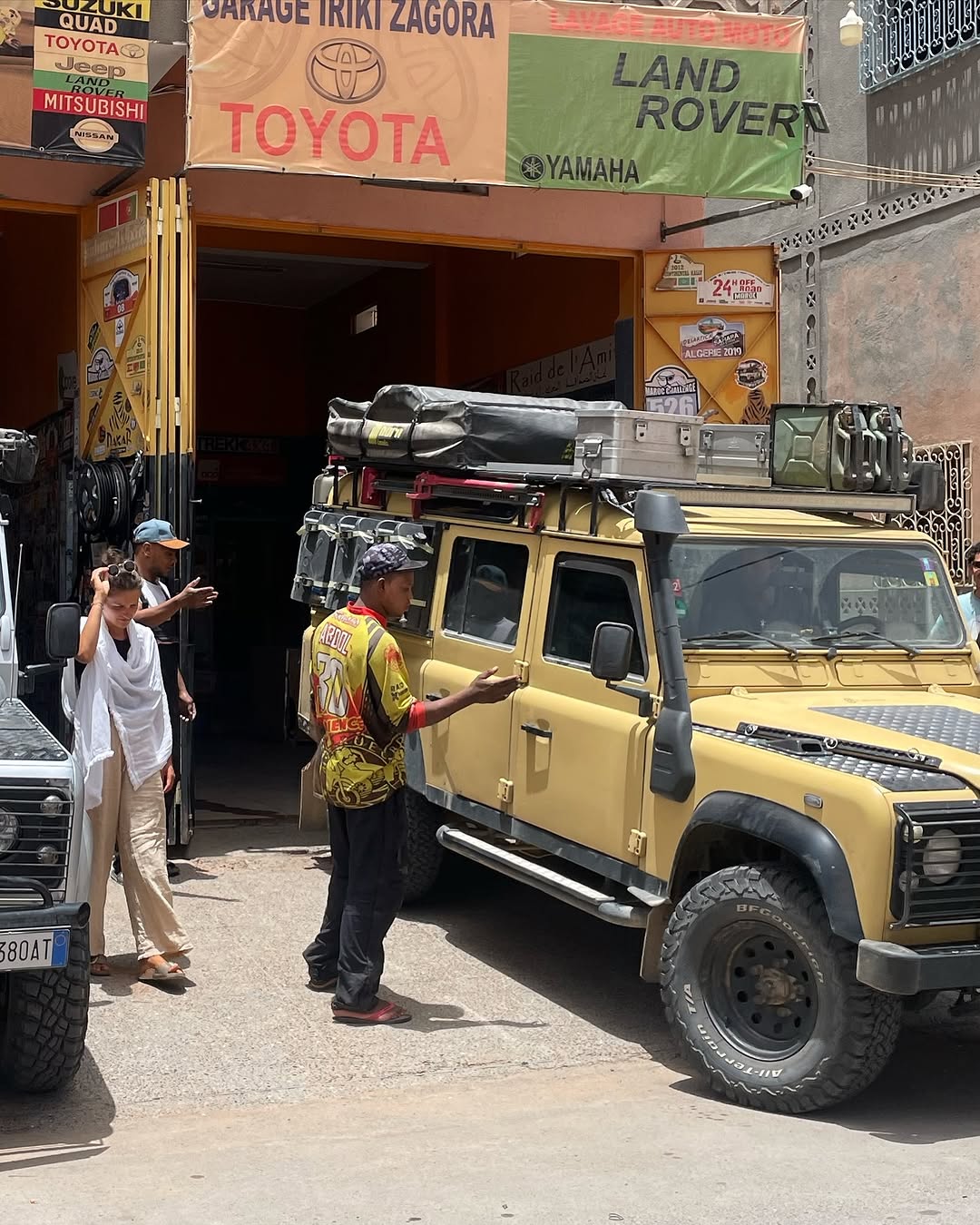 Sand-yellow Land Rover Defender 110 with German plates parked in front of Garage Iriki Zagora, Zakaria greeting the owners