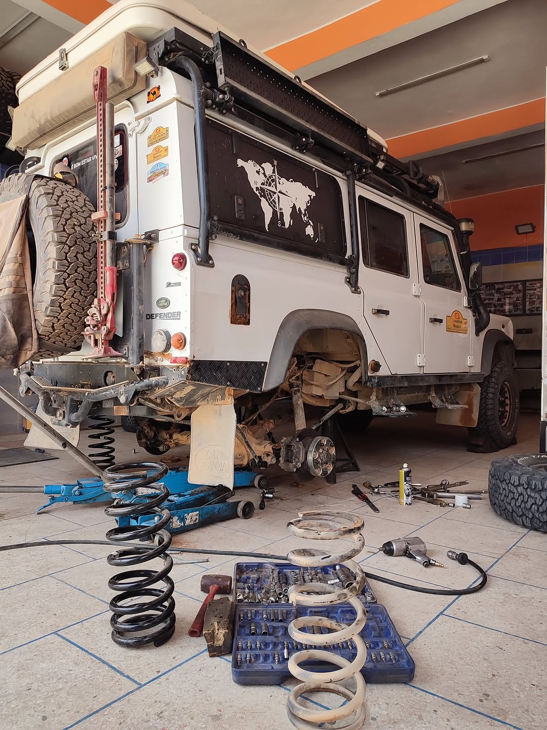 White Belgian Land Rover Defender 110 up on jacks at Garage Iriki, Zagora, with coil springs and tools spread across the floor during a rear suspension lift