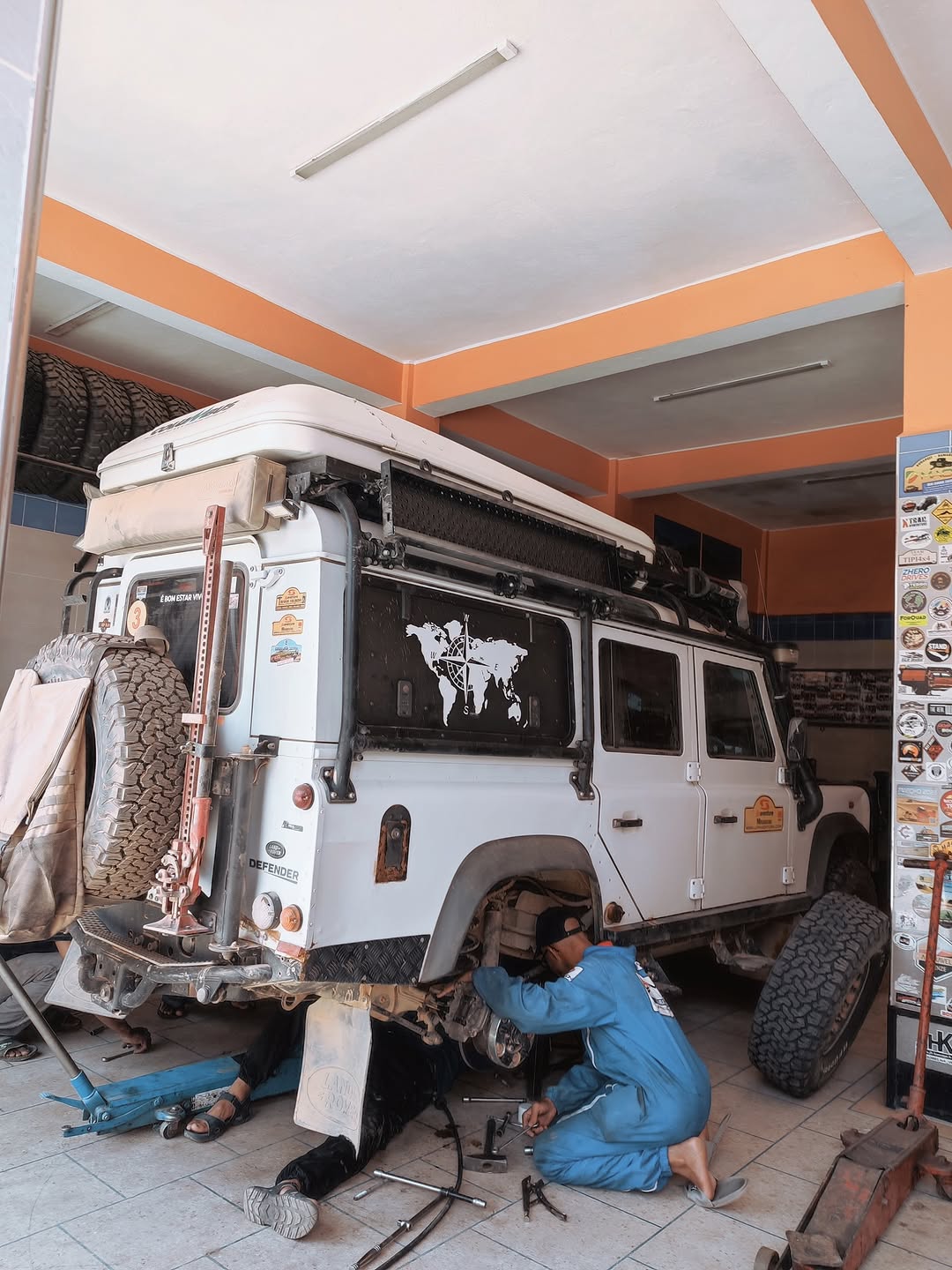Mechanic in blue overalls working under the raised Belgian Defender 110 at Garage Iriki, Zagora, refitting the reinforced rear suspension