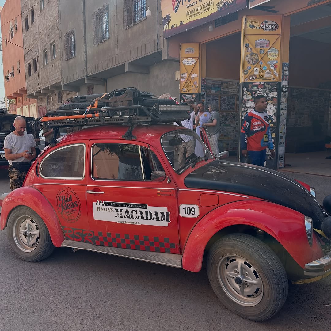 Side view of the red Rallye Macadam VW Beetle rally car with 'Bad Ideas, Good Stories' door decal outside Garage Iriki Zagora